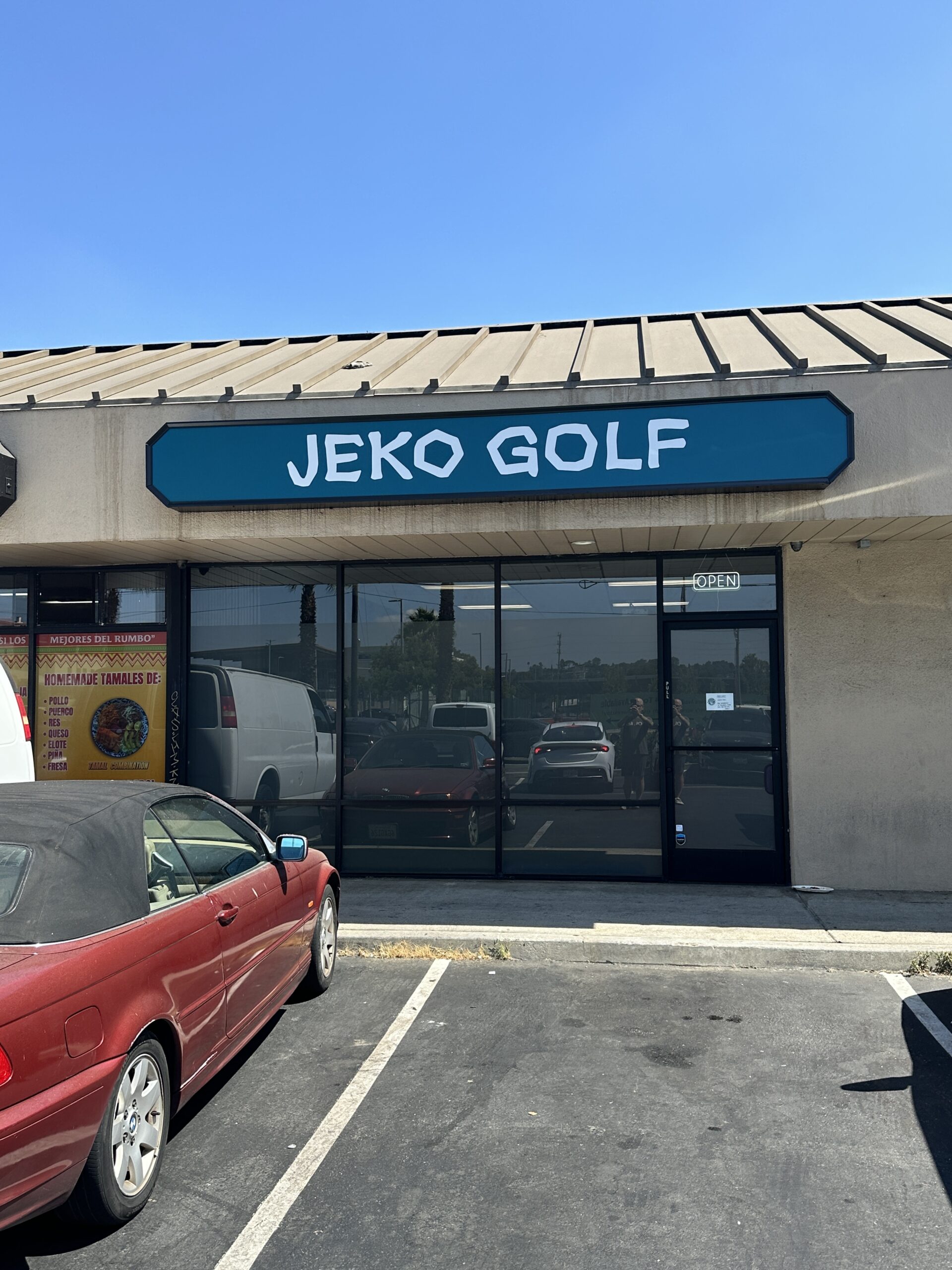 A photo of the Jeko Golf storefront. It's in a strip mall. Some cars are parked and reflected in the store's frosted windows. The sign above the door reads "Jeko Golf" and the sky is blue