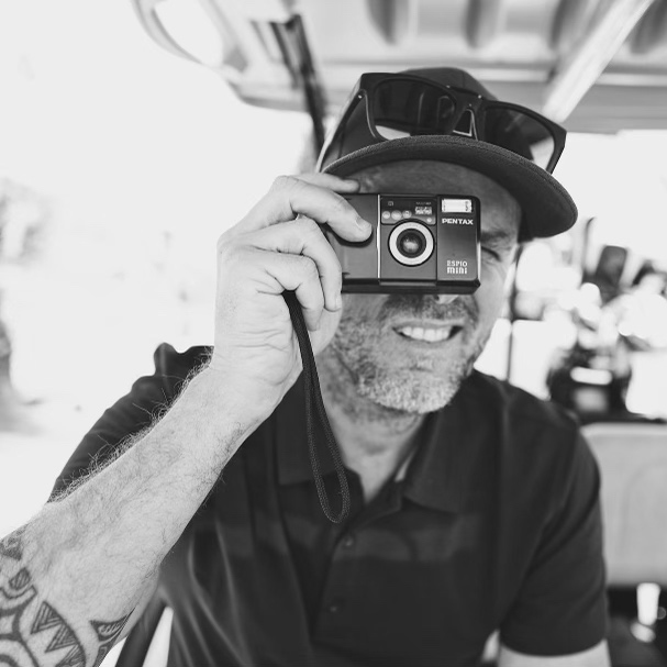 A photo of Josh, the owner of of Jeko Golf. It is in black and white and he his holding a small film camera. He is wearing a cap and sunglasses are resting atop the brim