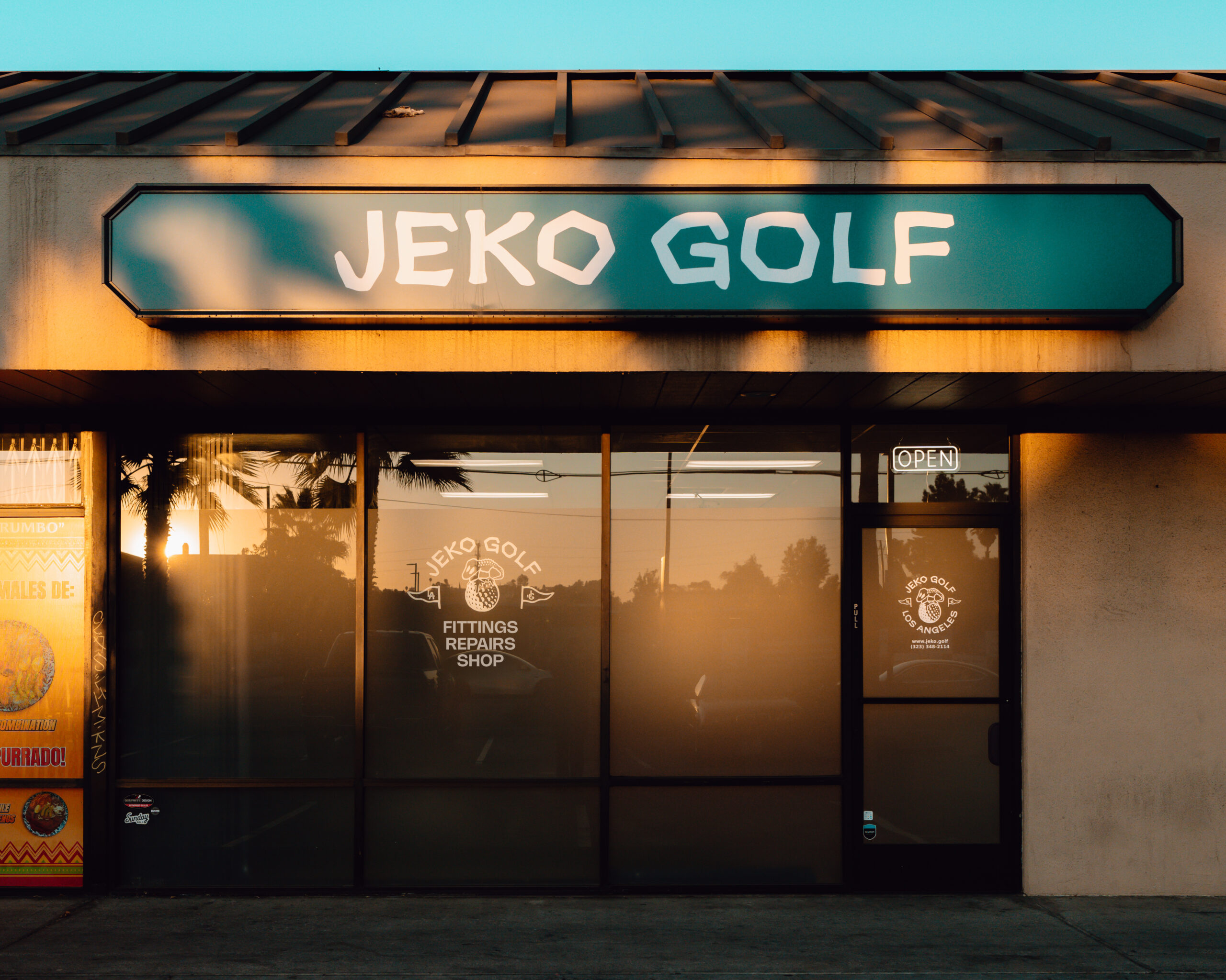 A photo of the storefront during the golden hour. The sunset is reflected in the shop's windows and palm tree shadows are cast onto the building.